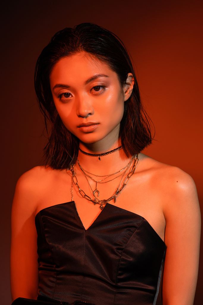 retrato de bela mulher asiática jovem com cabelo curto e penteado molhado posando em vestido preto sem alças, enquanto em pé sobre fundo laranja com iluminação vermelha, jóias douradas  - Foto, Imagem