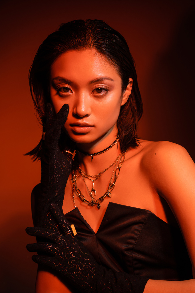 портрет потрясающей азиатской молодой женщины с короткими волосами и влажной прической, позирующей в черном платье без бретелек и перчатках, стоя на оранжевом фоне с красным освещением, золотыми украшениями  - Фото, изображение