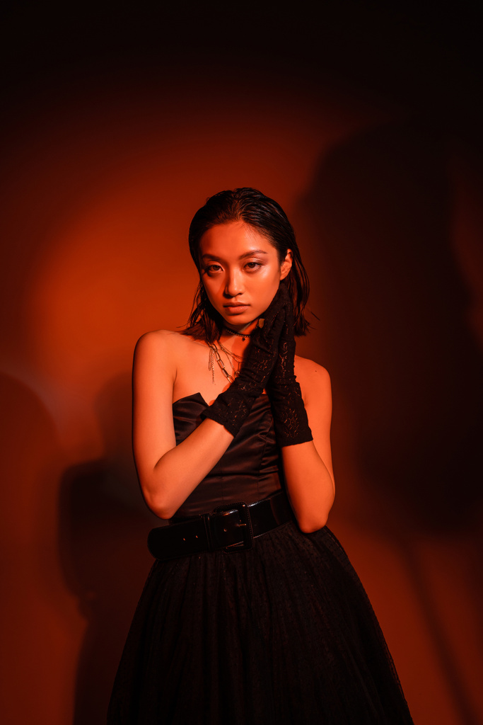 hezká asijská žena s krátkými vlasy a mokrý účes pózování v černé šaty bez ramínek s tylu sukně a rukavice při stání na tmavě oranžové pozadí s červeným osvětlením, zlaté šperky, mladý model - Fotografie, Obrázek