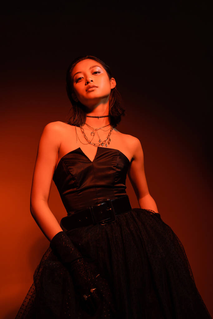 mujer asiática segura con peinado mojado posando en vestido negro sin tirantes con falda de tul y guantes mientras está de pie sobre fondo naranja oscuro con iluminación roja, joyas de oro, modelo joven - Foto, Imagen