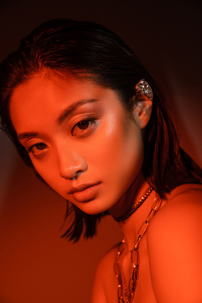 portret hipnotyzującej Azjatki z krótkimi włosami i mokrą fryzurą pozowanie z modnym kolczykiem mankietu i naszyjnikami na ciemnopomarańczowym tle z czerwonym oświetleniem, młoda modelka, patrząca w kamerę - Zdjęcie, obraz