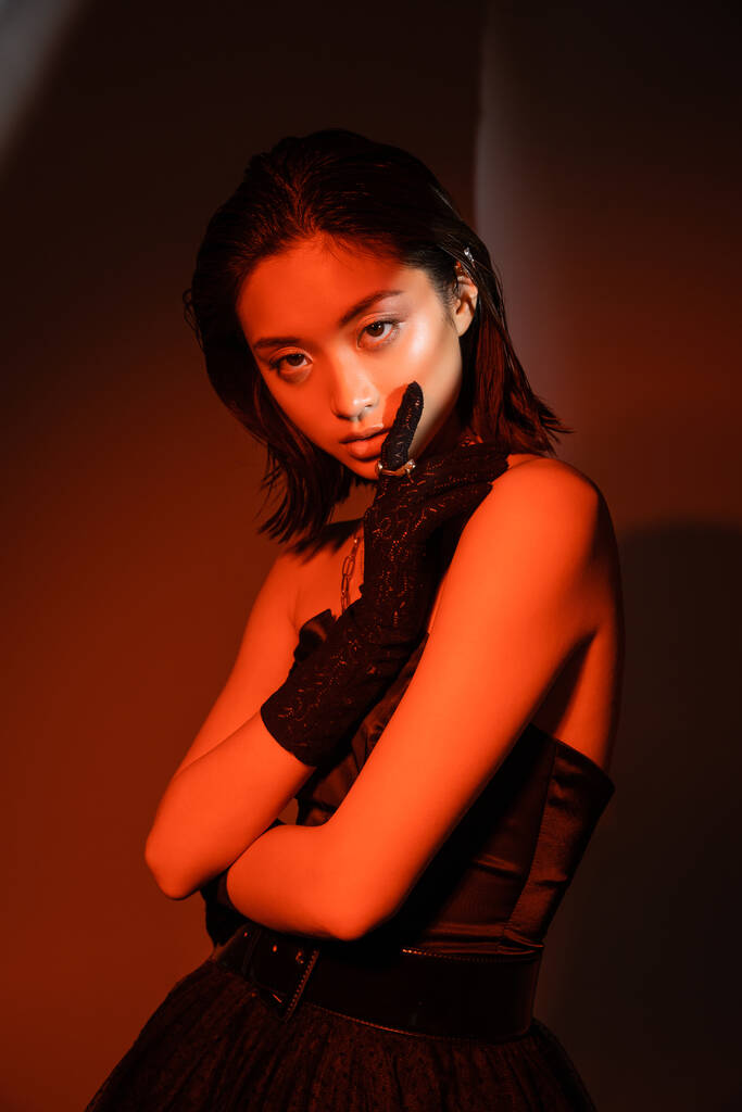 portrait of stunning asian woman with short hair and wet hairstyle posing in strapless dress and glove with golden rings with trendy cuff earring on orange background with red lighting, young model  - Photo, Image