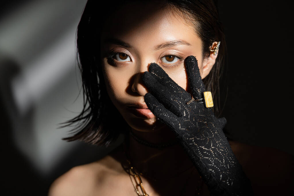 portrait of asian young woman with wet hairstyle and short hair in black glove with golden rings touching face while standing on grey background, model, looking at camera, shadows, dark - Photo, Image