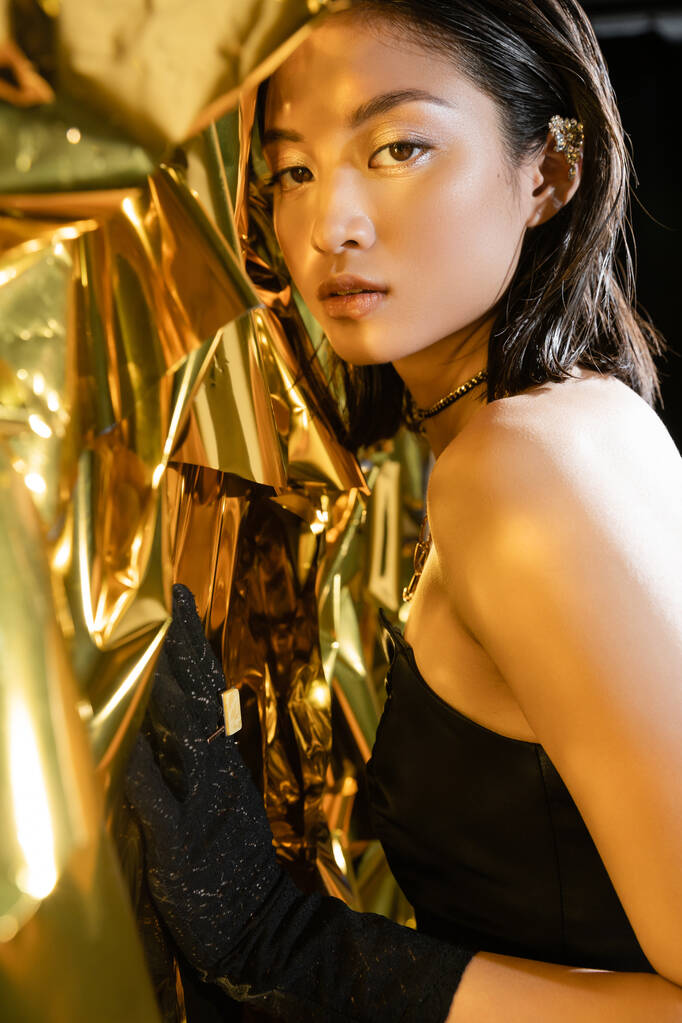 retrato de elegante mujer joven asiática con el pelo corto mojado posando en vestido negro sin tirantes junto al fondo amarillo brillante, modelo, mirando a la cámara, lámina dorada arrugada, belleza asiática natural  - Foto, imagen