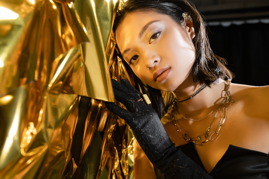 portrait de séduisante jeune femme asiatique aux cheveux courts mouillés et au gant noir touchant fond jaune brillant, mannequin, regardant la caméra, feuille d'or ridée, beauté asiatique naturelle  - Photo, image