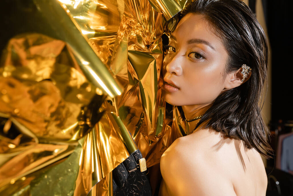 портрет привабливої азіатської молодої жінки з мокрим коротким волоссям, що позує поруч з блискучим золотим фоном, модель, дивлячись на камеру, зморщена жовта фольга, природна краса, чорна рукавичка
  - Фото, зображення
