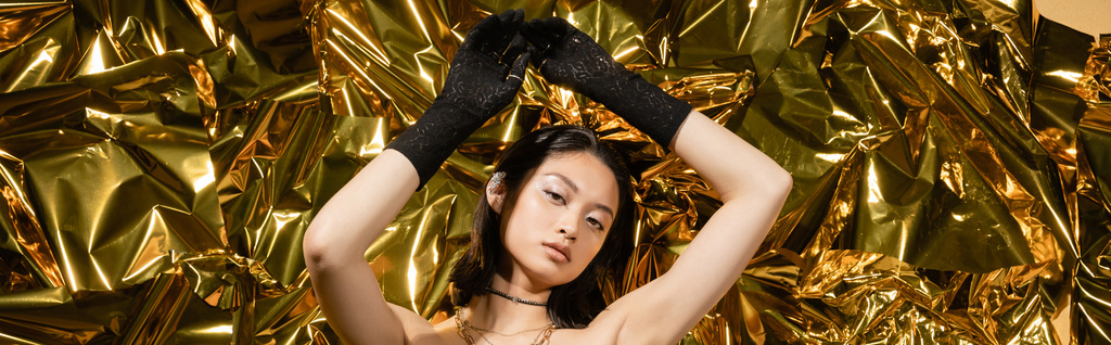 Islak saç stili ve kısa saçlı Asyalı genç kadın siyah eldivenlerle poz verirken parlak arka planının yanında duruyor, kameraya bakıyor, kırışık altın folyo, afiş  - Fotoğraf, Görsel
