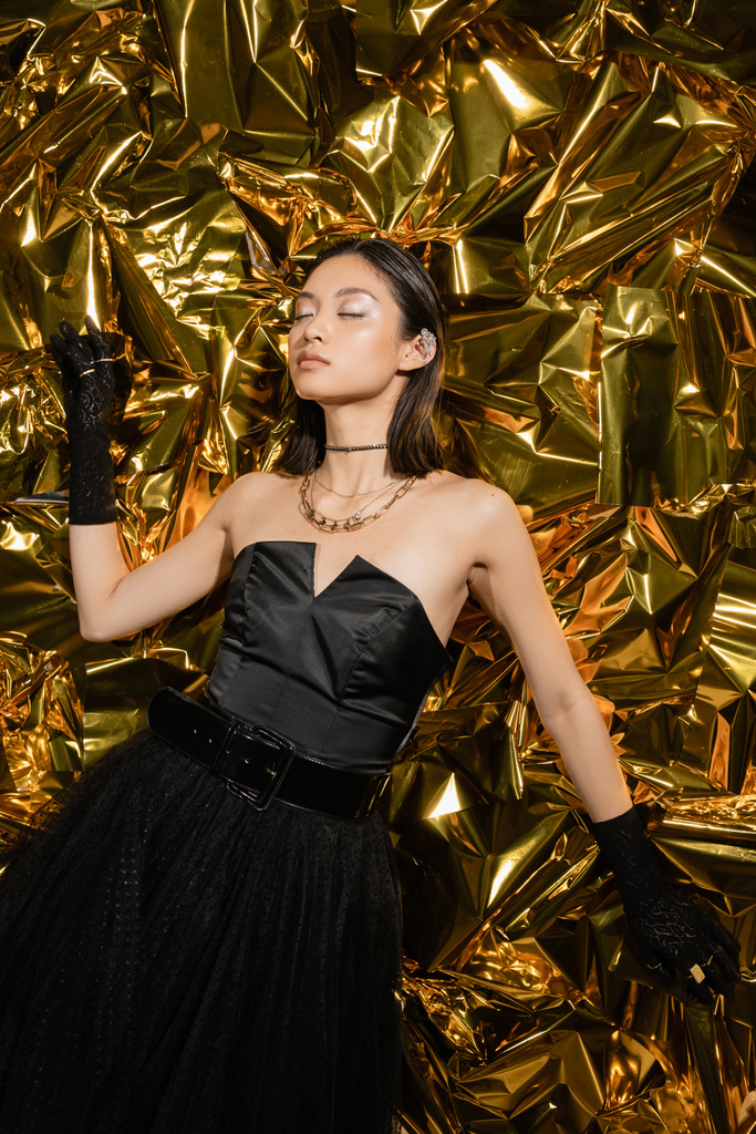 attractive asian young woman with short hair and closed eyes posing in black strapless dress with tulle skirt and gloves while standing next to shiny golden background, model, wrinkled foil - Photo, Image