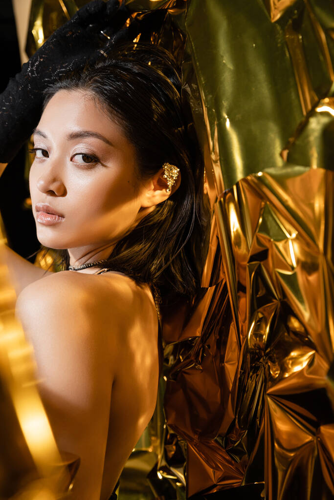 элегантная азиатская молодая женщина с влажной прической и короткими волосами, блестящими тенями для век, позирующая в черной перчатке, стоя рядом с блестящим фоном, модель, глядя в камеру, морщинистая золотая фольга - Фото, изображение