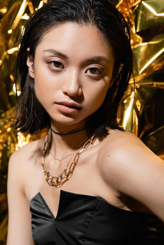 beautiful asian young woman with wet hairstyle and short hair posing in strapless dress with black glove while standing next to shiny golden background, model, looking at camera, wrinkled golden foil - Photo, Image