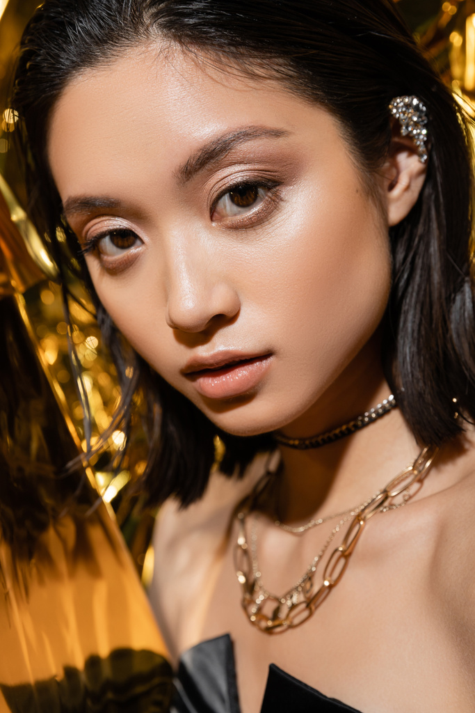 közelről csábító ázsiai fiatal nő nedves rövid haj pózol mellett fényes sárga háttér, modell, nézi a kamera, ráncos arany fólia, természetes smink, szépség  - Fotó, kép