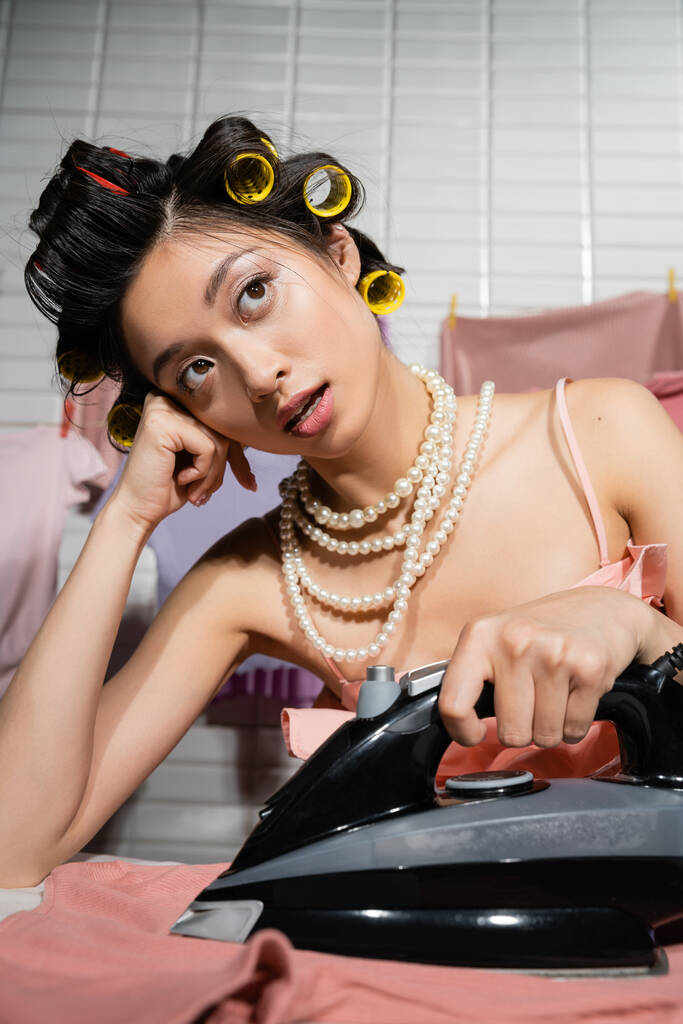 verträumte asiatische Hausfrau mit Haaren und Perlenketten-Bügeln und Wegschauen in der Nähe sauberer und nasser Kleidung, die auf verschwommenem Hintergrund hängt, Hausarbeit, junge Frau, Wäsche  - Foto, Bild
