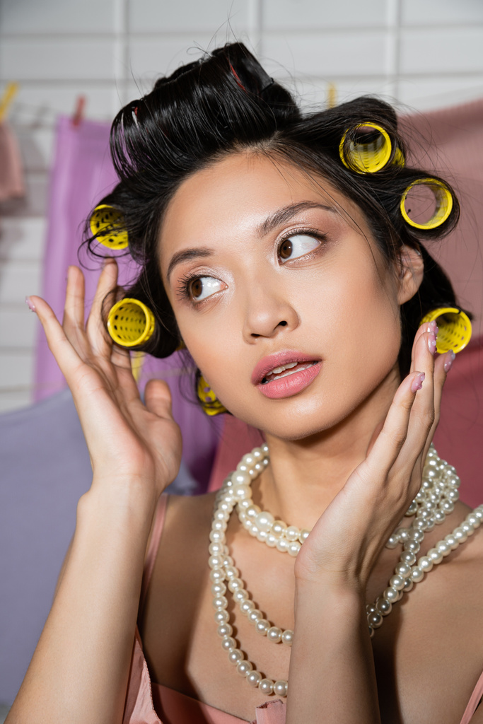 zamyšlená mladá asijská žena dotýkající se vlasů s vlasy natáčky a pózování v perleťovém náhrdelníku v blízkosti čisté a mokré prádelny visí na rozmazané pozadí, domácí práce, hospodyňky, dívá se jinam - Fotografie, Obrázek
