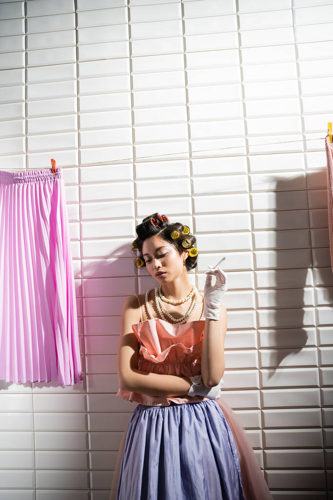 азиатская молодая женщина с бигуди волос стоя в розовом потрепанный топ, жемчужное ожерелье и перчатки, держа сигарету возле мокрой прачечной висит рядом с белой плиткой, домохозяйка, курение, плохая привычка  - Фото, изображение