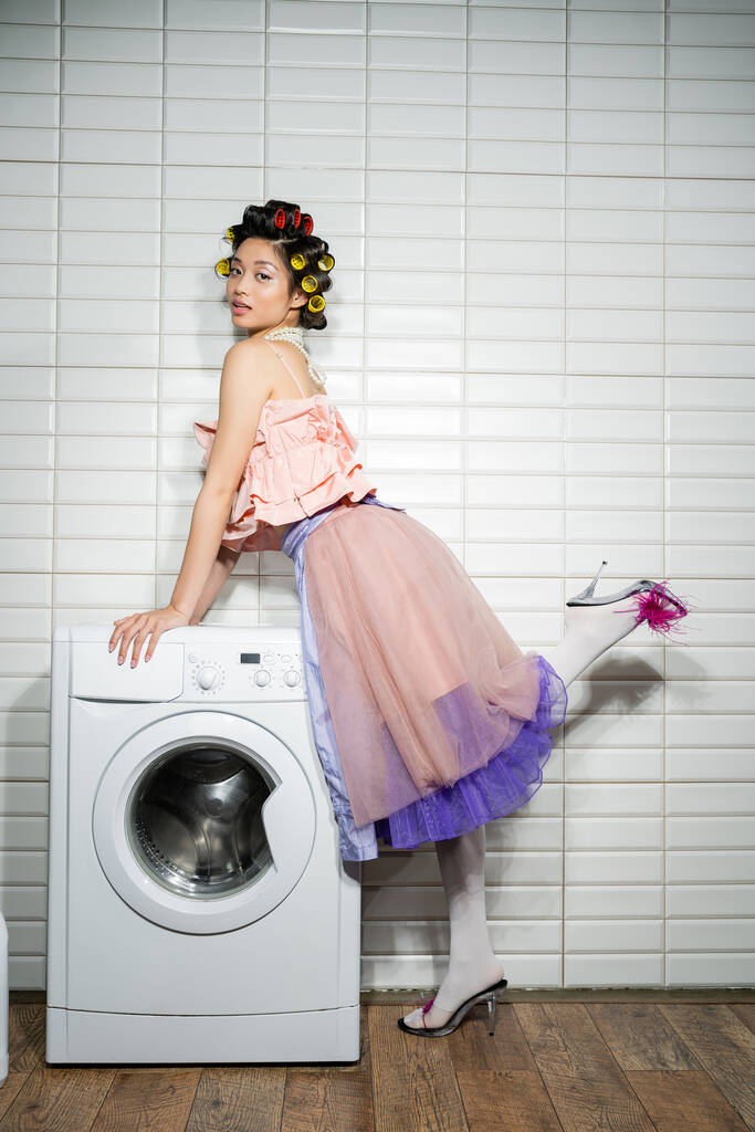 Pembe bukleli üst, inci kolye, tüy etek ve topuklu ayakkabı giyen Asyalı genç kadın çamaşır odasındaki beyaz fayansların yanında modern çamaşır makinesinin yanında duruyor.  - Fotoğraf, Görsel