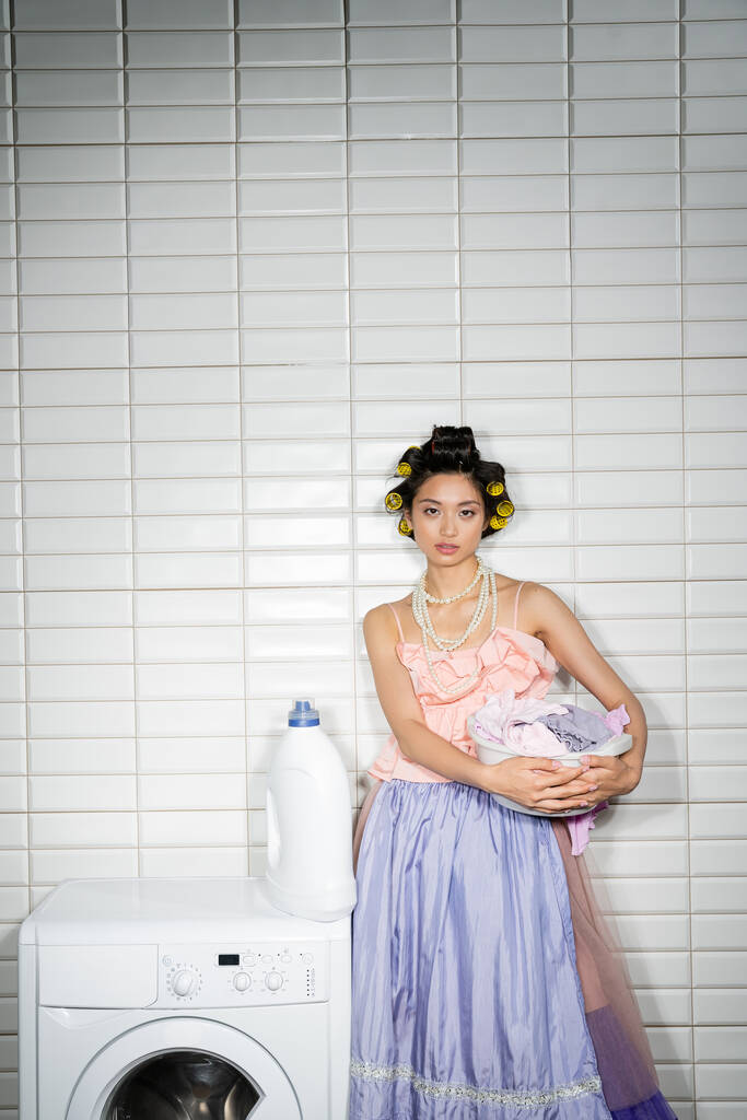 aziatische jonge vrouw met haar krulspelden staan in roze ruches top, parel ketting en tule rok en houden waskom met vuile kleren in de buurt van moderne wasmachine en wasmiddel in wasruimte  - Foto, afbeelding