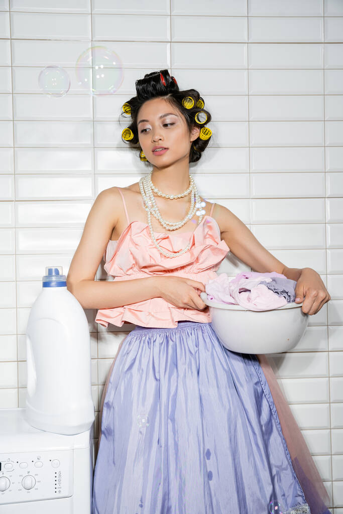 aziatische jonge vrouw met haar krulspelden staan in roze ruches top, parel ketting, tule rok en houden waskom met vuile kleren in de buurt van wasmachine en wasmiddel in wasruimte, zeepbellen - Foto, afbeelding