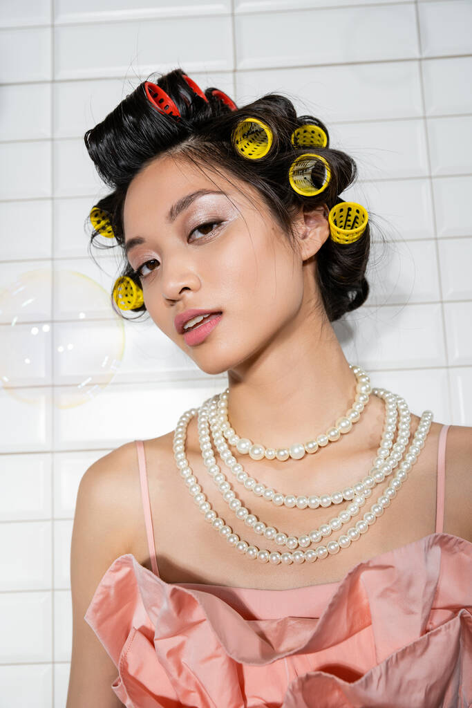 портрет брюнетки и молодой азиатской женщины с бигуди волос, стоящих в жемчужном ожерелье возле размытого мыльного пузыря в прачечной с белой плиткой, домохозяйка, природная красота  - Фото, изображение
