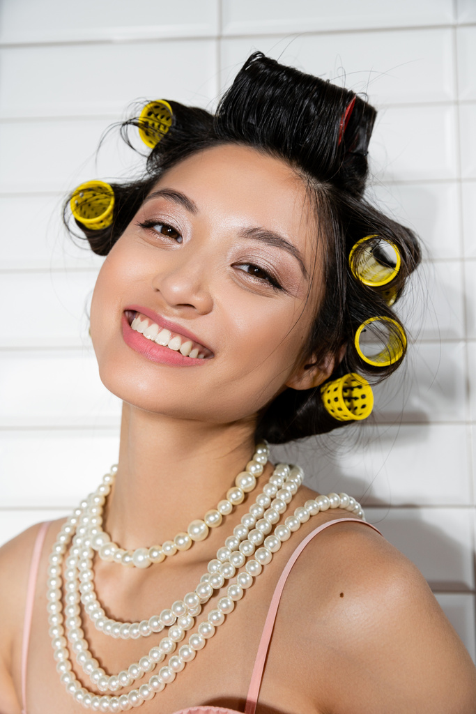 retrato de alegre e jovem mulher asiática com encrespadores de cabelo em pé no colar de pérolas e sorrindo na lavanderia com azulejos brancos, dona de casa, beleza natural  - Foto, Imagem