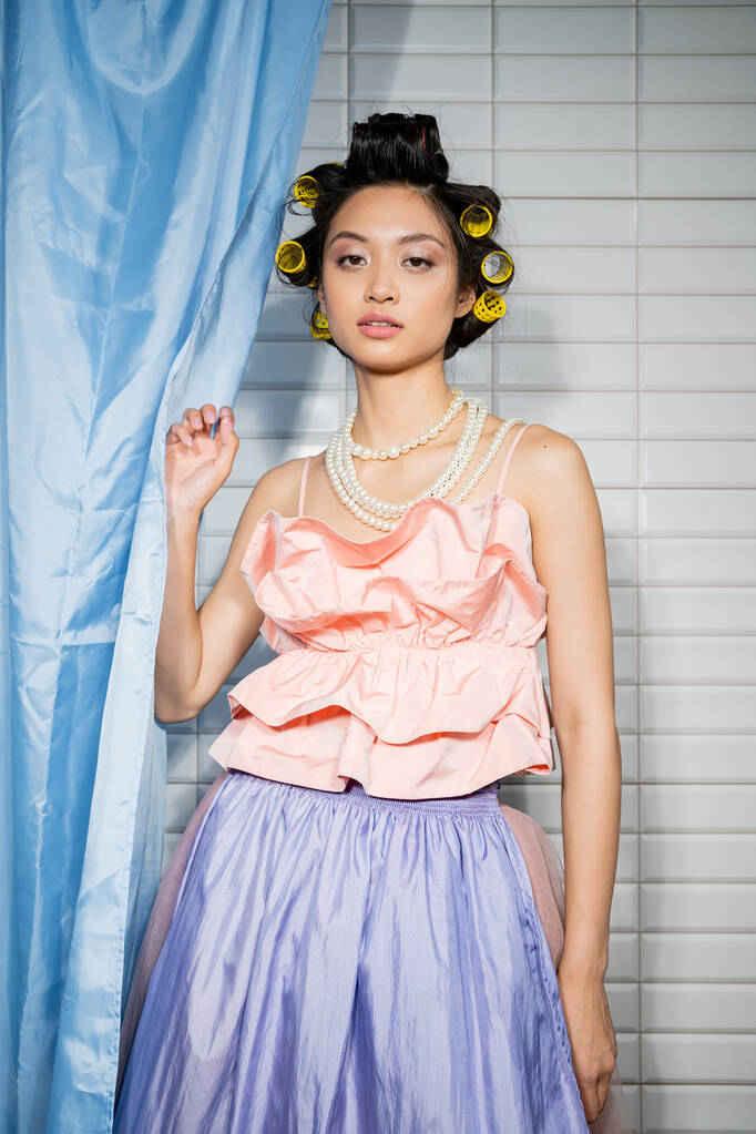 jeune femme à la mode et asiatique avec des boucles de cheveux debout en haut à volants rose avec collier de perles près du rideau de salle de bain bleu et en regardant la caméra près de carreaux blancs à la maison  - Photo, image