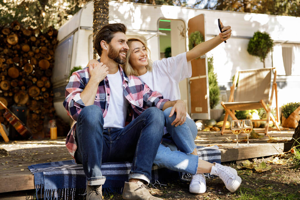 Ein nettes Paar sitzt neben dem Reiseanhänger, macht ein Selfie, lacht, genießt seinen Urlaub. Porträt einer Verliebten, die einen sonnigen Tag außerhalb der Stadt genießt und Wein trinkt - Foto, Bild