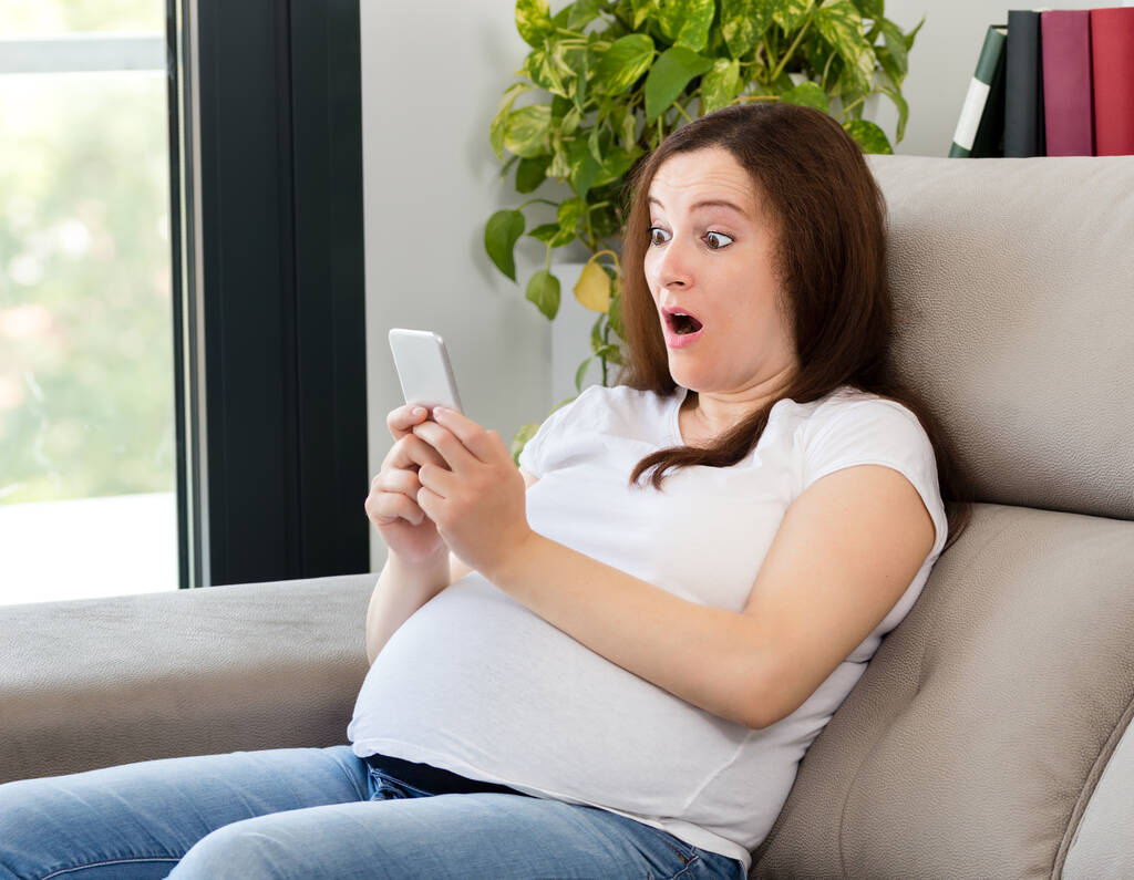 Έκπληκτη έγκυος γυναίκα κοιτάζοντας έξυπνο τηλέφωνο στο σπίτι - Φωτογραφία, εικόνα