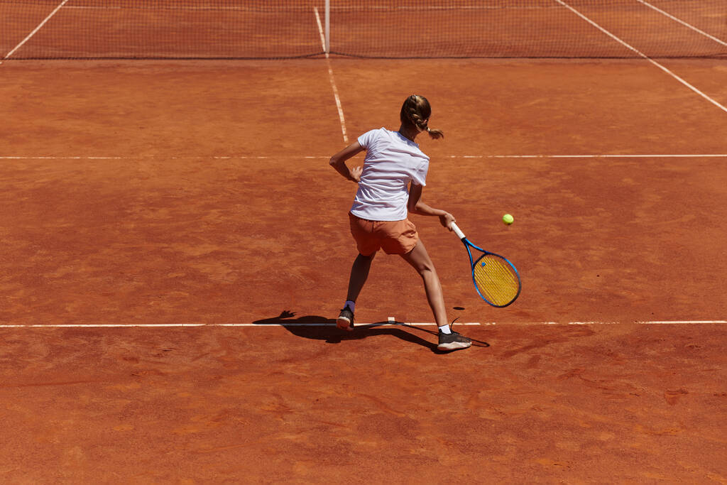 Ένα νεαρό κορίτσι που δείχνει επαγγελματικές δεξιότητες τένις σε ένα ανταγωνιστικό αγώνα σε μια ηλιόλουστη μέρα, που περιβάλλεται από τη σύγχρονη αισθητική ενός γηπέδου τένις - Φωτογραφία, εικόνα