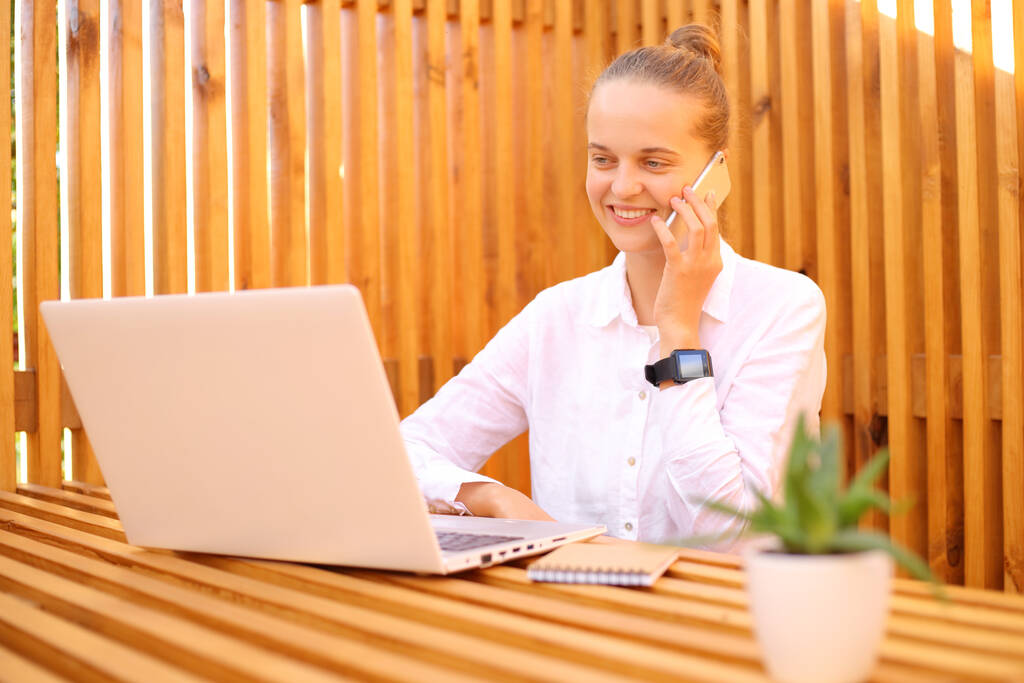 Усміхнена весела жінка фрілансер в білій сорочці сидить у відкритому кафе, працює на ноутбуці та розмовляє на смартфоні, ділова розмова з партнером
. - Фото, зображення