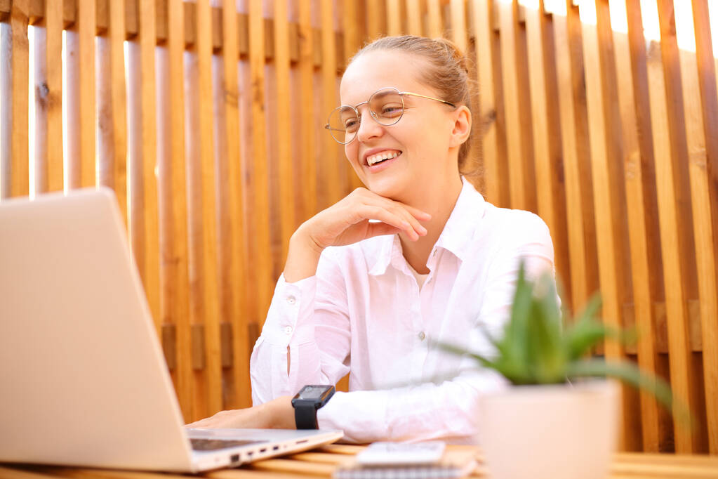 Zufrieden lächelnde Frau in weißem Hemd sitzt im Outdoor-Café und arbeitet am Laptop, während sie mit ihrem Geschäftspartner Videokonferenzen abhält. - Foto, Bild