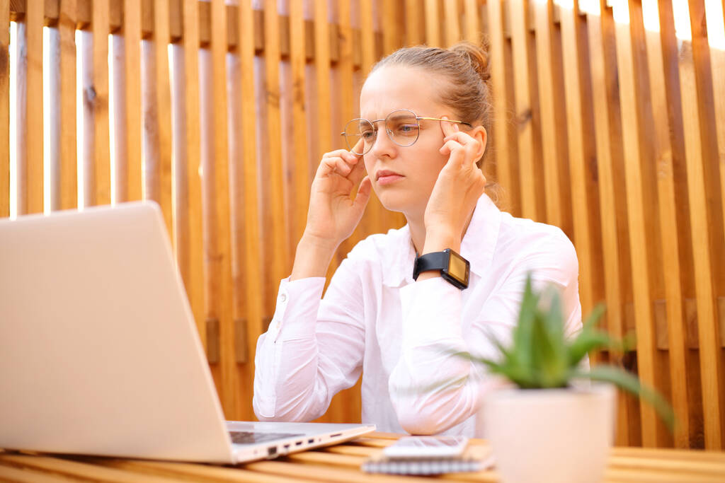 Väsynyt keskittynyt nainen valkoisessa paidassa istuu ulkona kahvilassa työskentelee kannettavan tietokoneen hierova temppeleitä on päänsärky ajatellut uusia työtehtäviä. - Valokuva, kuva