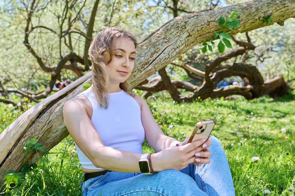 Mujer joven sentada en el césped del parque, relajándose con un smartphone. Aplicaciones móviles para ocio, aprendizaje, compras, relajación, tecnología de Internet en línea - Foto, Imagen