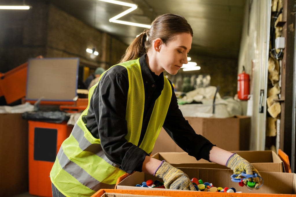 Young female worker of garbage sorting center wearing protective clothing and gloves while working with plastic caps in carton boxes, garbage sorting and recycling concept - Photo, Image