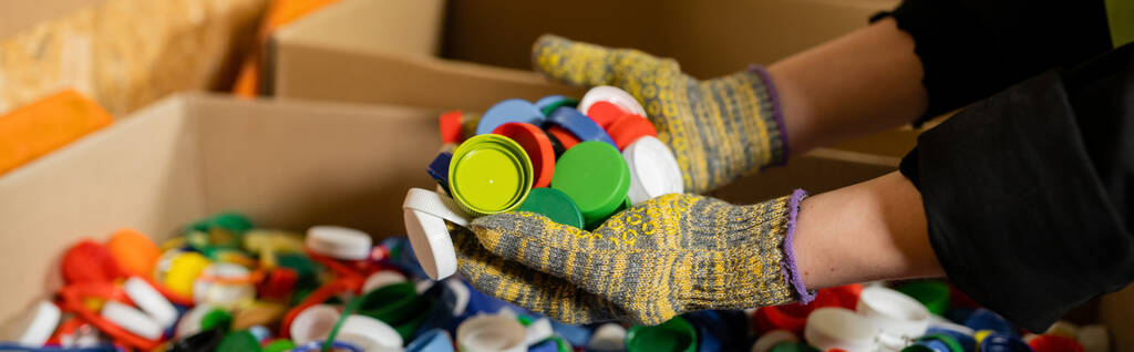 Widok przyciętych żeńskich sortowników w rękawiczkach ochronnych z kolorowymi plastikowymi wieczkami w pobliżu zamazanych pudełek kartonowych podczas pracy na stanowisku utylizacji odpadów, koncepcja sortowania i recyklingu śmieci, baner  - Zdjęcie, obraz