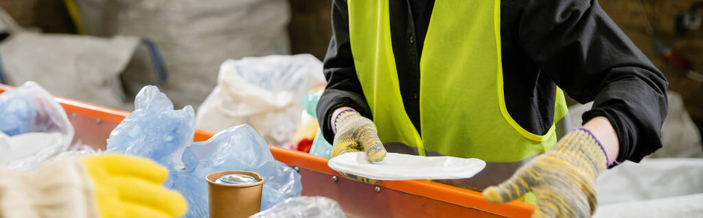 Gesneden weergave van sorteerder in beschermvest en handschoenen die vuilnis van transportband meenemen tijdens het werken in afvalverwerkingsstation op de achtergrond, vuilnissorteer- en recyclingconcept, banner  - Foto, afbeelding