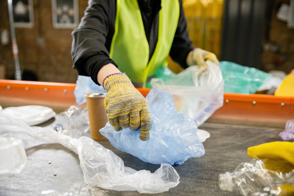 Rajattu näkymä hämärtynyt työntekijä suojaava liivi ja käsineet ottaen muovipussi kuljettimesta työskennellessään jäteasema taustalla, roskien lajittelu käsite  - Valokuva, kuva
