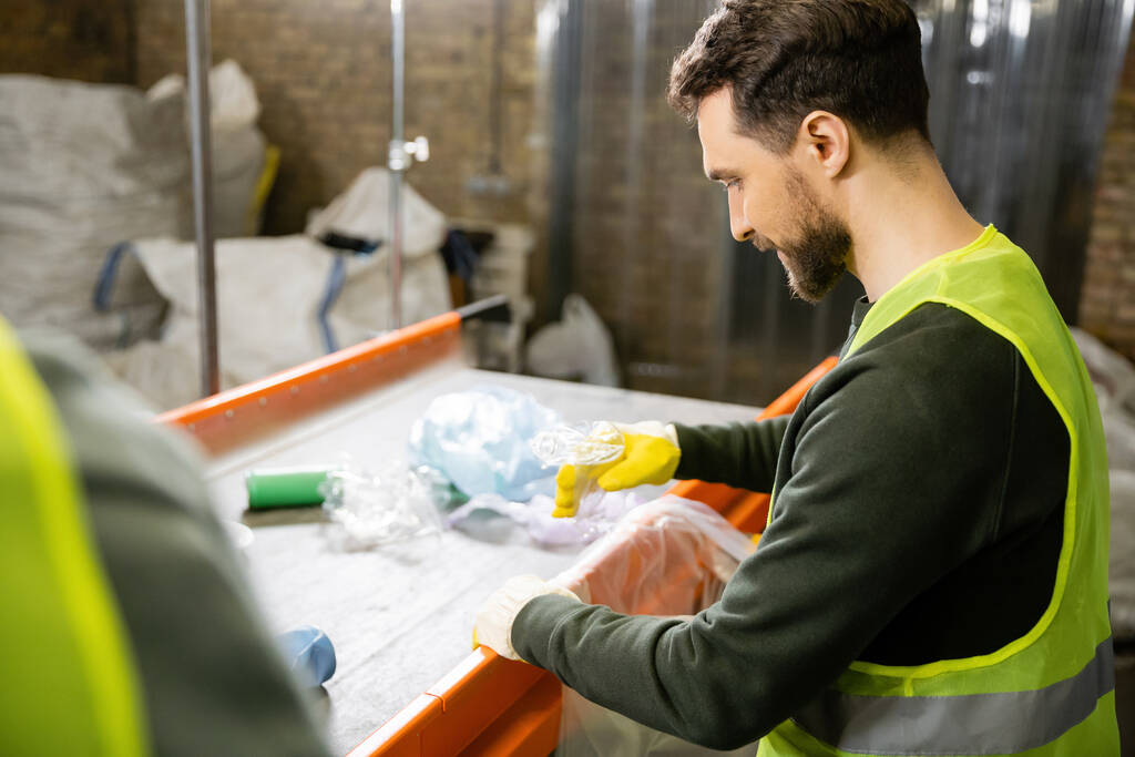 Side view of worker in protective gloves and vest sorting garbage near conveyor while standing in blurred waste disposal station at background, garbage sorting and recycling concept - Photo, Image
