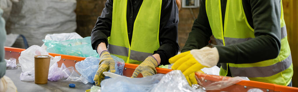 Cropped view of sorters in high visibility vests and gloves sorting garbage on conveyor while working in waste disposal station at background, garbage sorting and recycling concept, banner  - Photo, Image