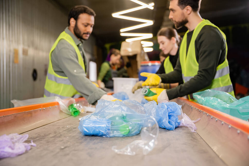Plastic trash on conveyor near blurred multiethnic workers in high visibility vests and gloves working together in garbage sorting center, garbage sorting and recycling concept - Photo, Image