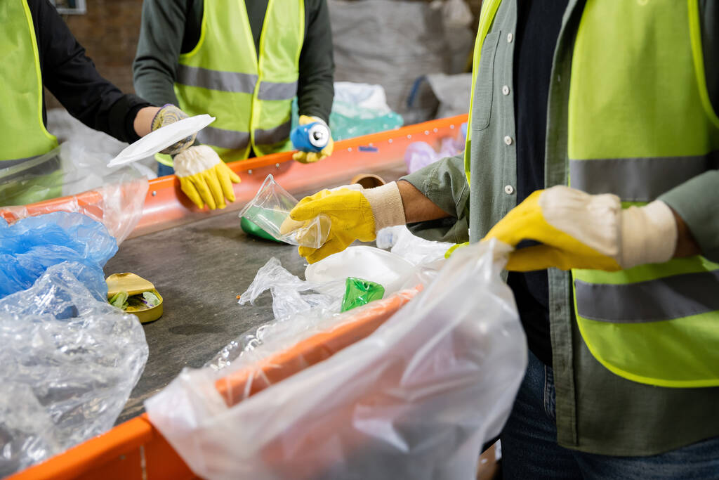 Gesneden weergave van sorteerder in handschoenen en beschermvest met wazig plastic zak en het nemen van afval van transportband tijdens het werken in de buurt van collega 's in afvalverwerkingsstation, afvalrecycling concept - Foto, afbeelding