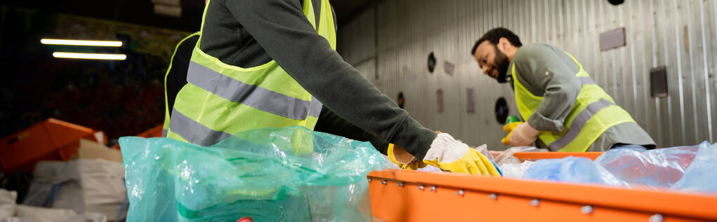 Άνδρας εργαζόμενος σε προστατευτικά γάντια και ανακλαστικό γιλέκο λαμβάνοντας σκουπίδια από μεταφορέα κοντά σε πλαστική σακούλα και θολή ινδική συνάδελφο στο παρασκήνιο στο σταθμό διάθεσης αποβλήτων, έννοια ανακύκλωσης, πανό  - Φωτογραφία, εικόνα