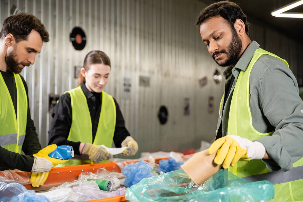 Bearded indian worker in safety vest and protective gloves putting paper cup in plastic bag while separating trash near conveyor and colleagues in waste disposal station, recycling concept - Photo, Image