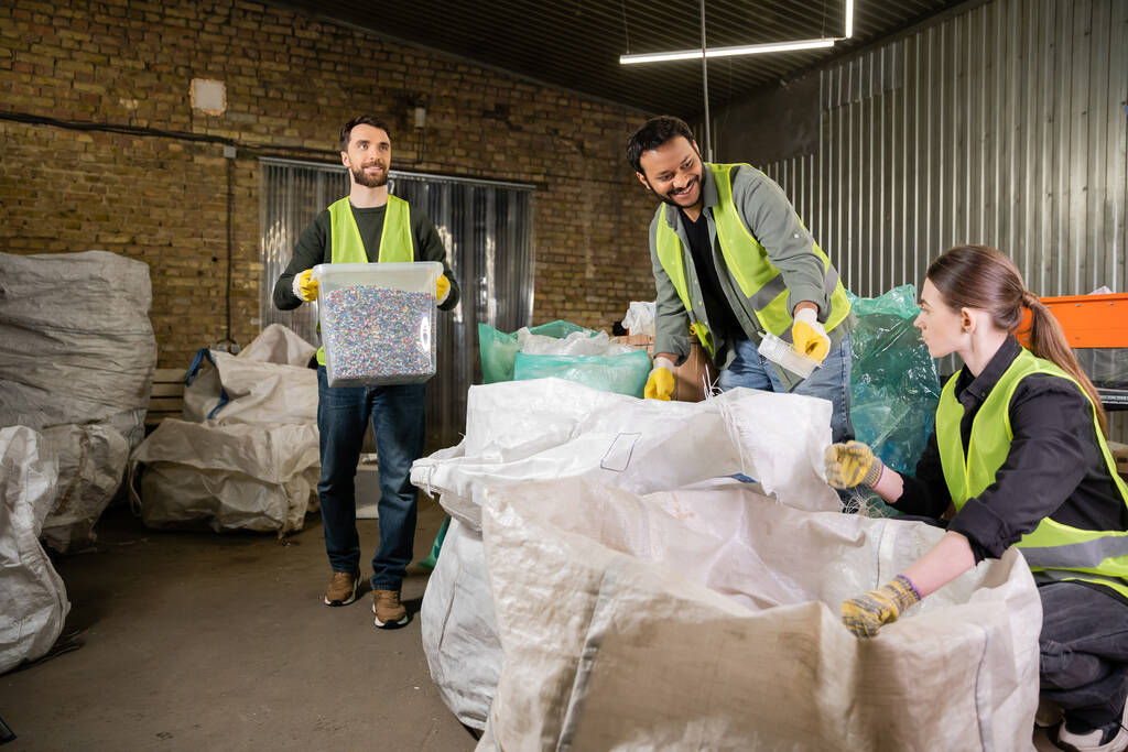Glimlachende Indiase werknemer in veiligheidsvest en handschoenen geeft plastic afval aan collega in de buurt van zakken en sorteerder met container tijdens het werken in afvalverwerkingsstation, recycling concept - Foto, afbeelding