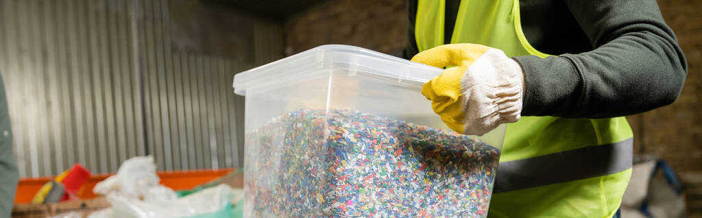 Widok sortownika w kamizelce ochronnej i rękawicy trzymającej pojemnik z tworzywa sztucznego do recyklingu podczas pracy w rozmazanym centrum sortowania śmieci, sortowania śmieci i recyklingu koncepcji, baner  - Zdjęcie, obraz