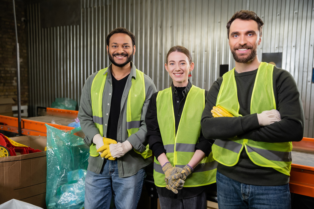 Positive interracial workers in high visibility vests and gloves looking at camera while standing near conveyor and working in waste disposal station, recycling concept - Photo, Image