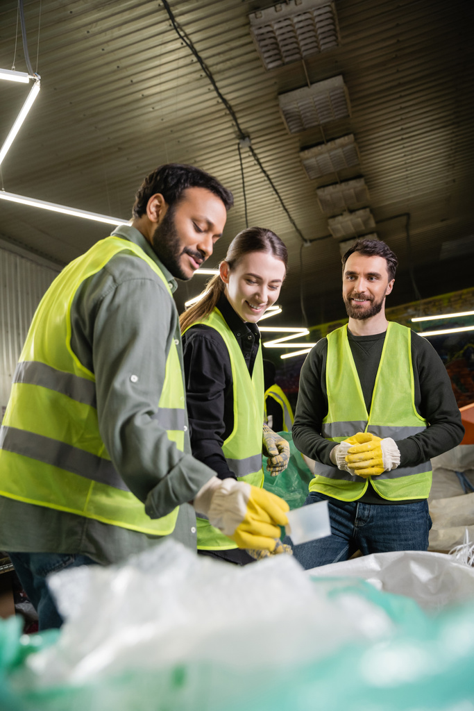 Fröhlicher junger Arbeiter in Warnweste und Handschuhen, der neben multiethnischen Kollegen in der Nähe von Säcken in einer verschwommenen Müllentsorgungsstation steht  - Foto, Bild