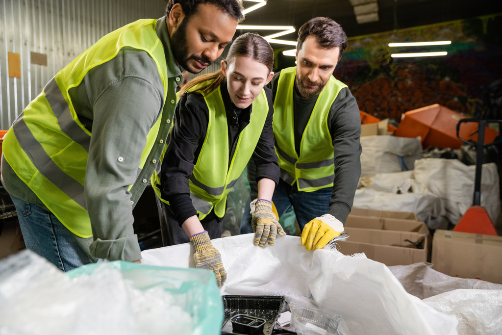 Jovem trabalhadora em jaqueta de alta visibilidade e luvas olhando para sacos enquanto trabalhava em conjunto com colegas masculinos multiétnicos em estação de descarte de resíduos desfocada, processo de triagem de lixo  - Foto, Imagem