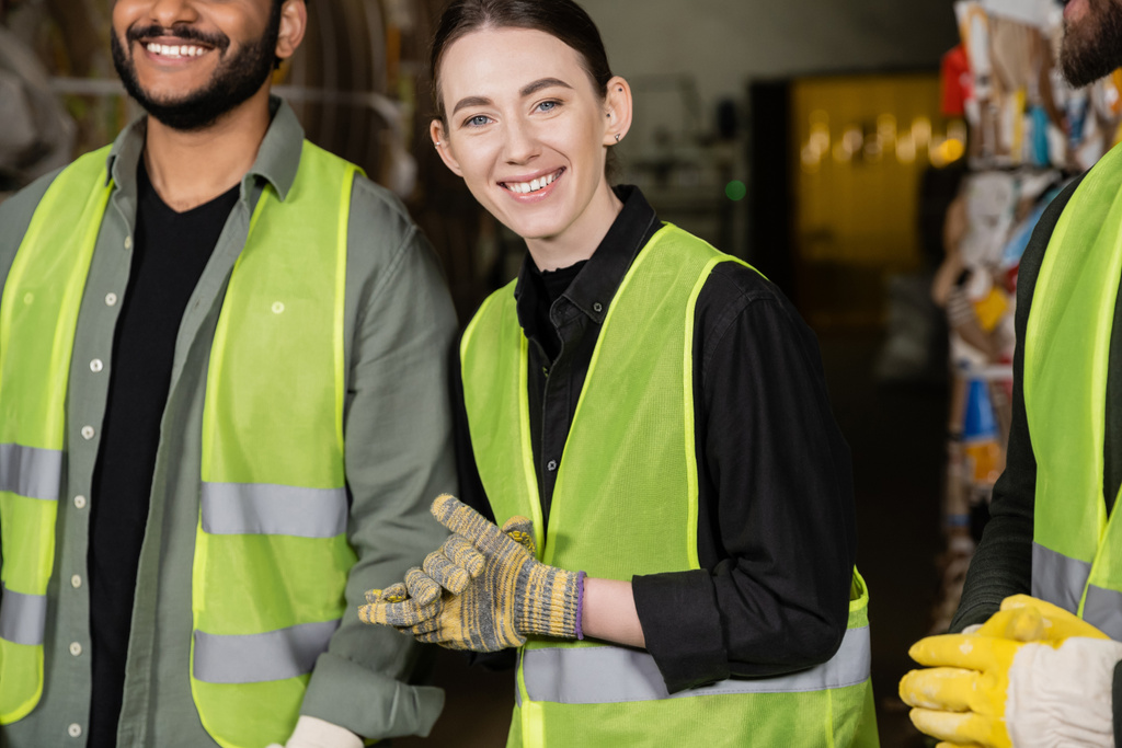 Young worker in high visibility vest and gloves smiling at camera while standing near multiethnic colleagues in blurred waste disposal station, garbage sorting process  - Photo, Image