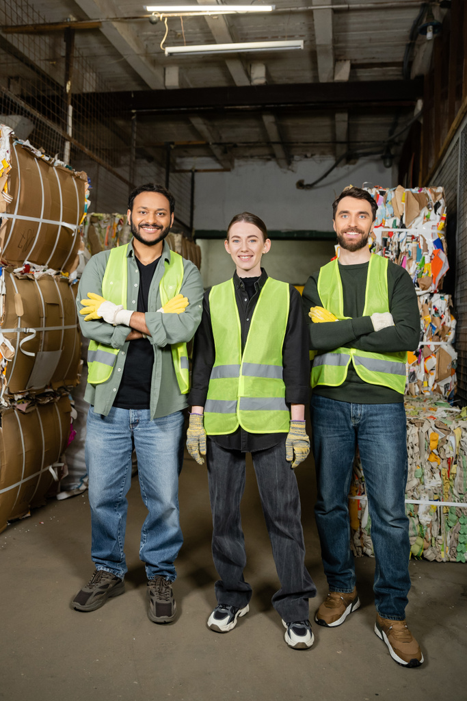 Smiling and interracial workers in high visibility vests and gloves crossing arms and looking at camera while standing near waste paper on disposal station, garbage sorting process  - Photo, Image