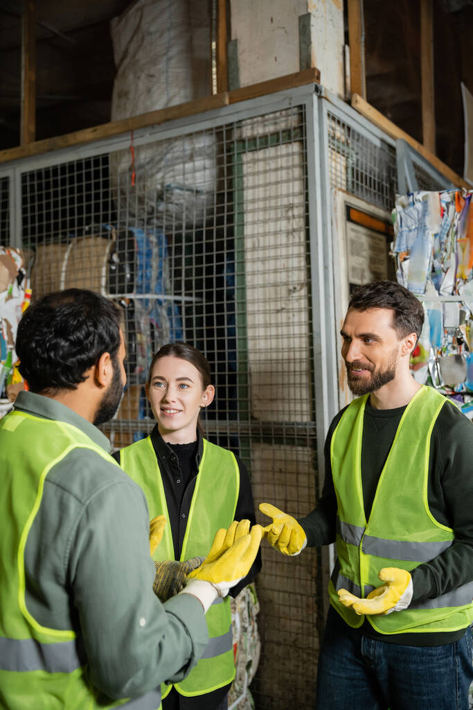Smiling male worker in safety vest and gloves talking to indian colleague while resting near waste paper in garbage sorting center, waste sorting and recycling concept - Photo, Image