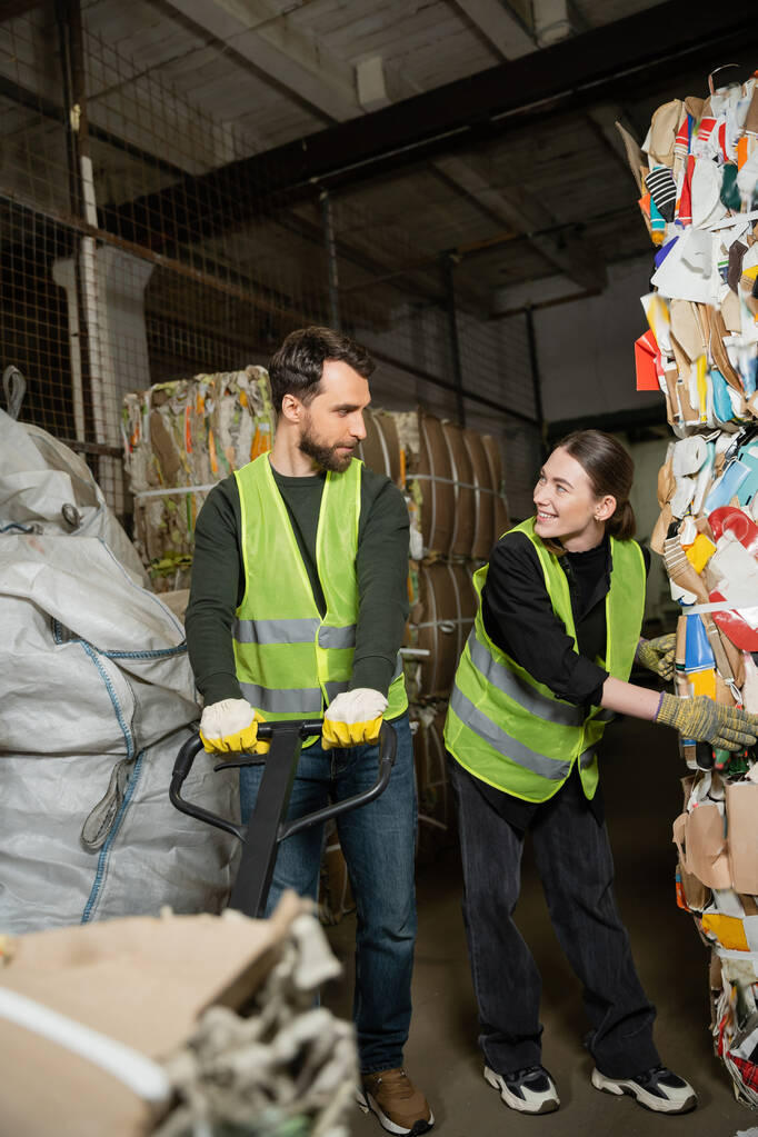 Smiling woman in high visibility vest and gloves talking to male colleague with hand pallet truck while working with waste paper in blurred garbage sorting center, waste sorting and recycling concept - Photo, Image